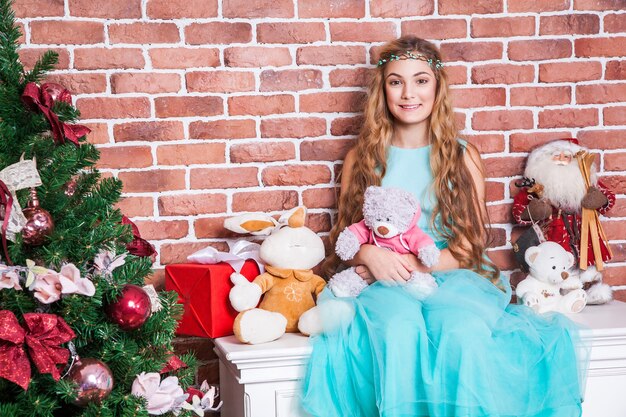 Atractiva rubia de pelo largo adolescente sentada en una mesita de noche blanca cerca del árbol de Navidad, sonrisa con dientes y mirando a cámara. Tiro del estudio