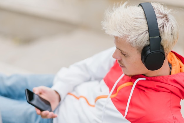 Foto atractiva vista superior de un joven rubio disfrutando del clima en el parque de la ciudad esperando el mensaje de texto de su novia mientras escucha su canción favorita usando el teléfono móvil al aire libre en el parque