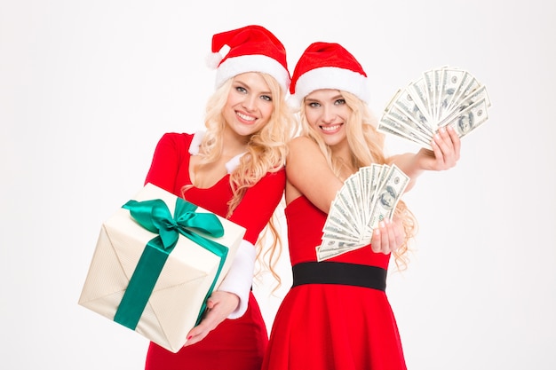 Atractivas hermanas gemelas rubias alegres en vestidos y sombreros rojos de santa claus posando con dinero y presente sobre fondo blanco.