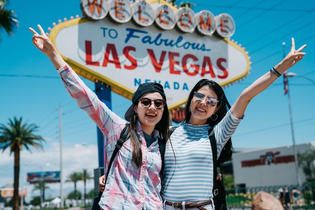 Atractivas y hermosas compañeras de viaje con sonrisas radiantes y posando con el dedo de la victoria afuera. mochileros femeninos emocionados en un día soleado tomando una cámara de fotos con la cartelera de las veags.