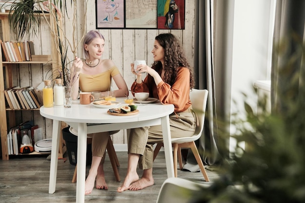 Atractivas jóvenes lesbianas sentadas en la mesa del comedor y tomando café mientras conversan durante el desayuno en casa