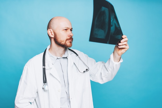 Atractivo amable médico barbudo calvo mirando la radiografía aislada sobre fondo azul.