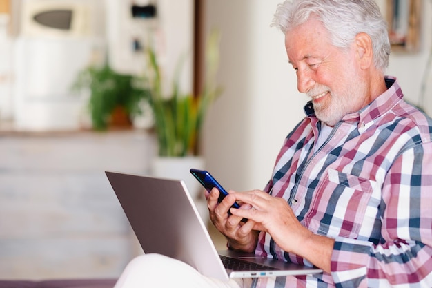 Atractivo anciano navegando por una computadora portátil sosteniendo un teléfono móvil Abuelo anciano alegre y sonriente en casa estando de buen humor navegando por la red