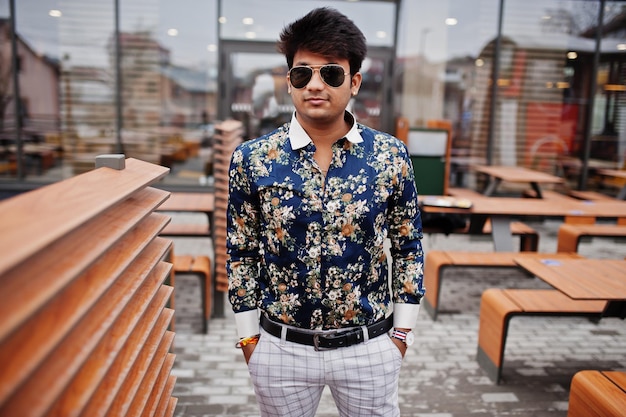 Atractivo y apuesto hombre indio elegante vestido con camisa y gafas de sol parado en la terraza del moderno café salón en su descanso de trabajo