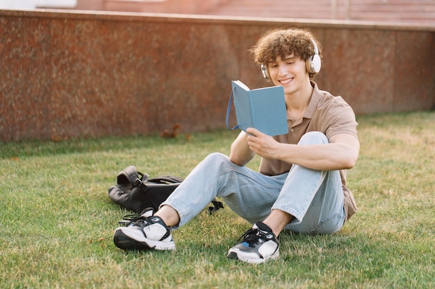 Atractivo estudiante masculino en auriculares estudiando cerca de la universidad sentado en el césped preparándose para los exámenes