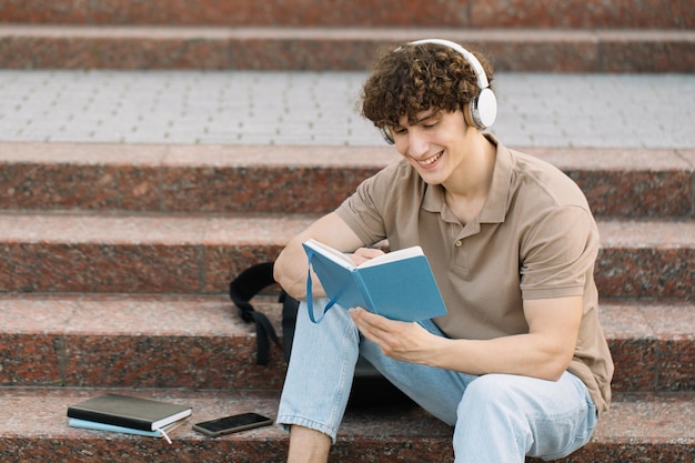 Atractivo estudiante masculino se sienta en los escalones de la universidad y lee algo preparándose para los exámenes