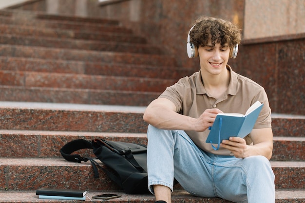 Atractivo estudiante masculino se sienta en los escalones de la universidad y lee algo preparándose para los exámenes