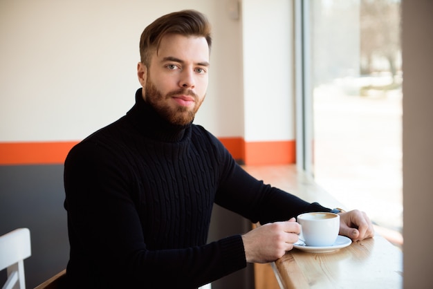 Atractivo exitoso joven empresario beber café sentado en la mesa de café.