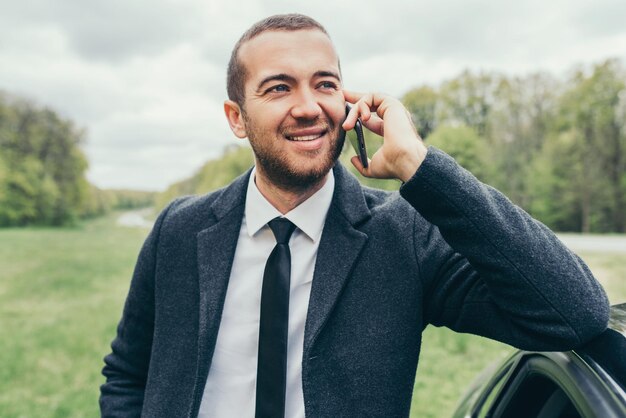 Atractivo gerente caucásico europeo con un elegante abrigo formal que usa un teléfono móvil cerca de su auto mientras espera que sus colegas prueben la conducción Elegante hombre de negocios sonriente y feliz tiene una charla en un teléfono inteligente