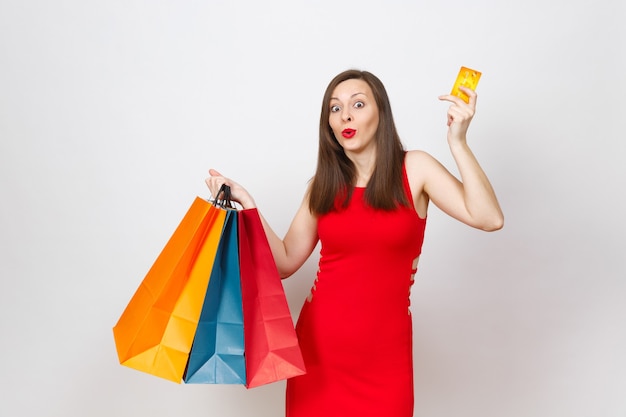 Atractivo glamour moda joven mujer de cabello castaño en vestido rojo con tarjeta de crédito, paquetes multicolores con compras después de ir de compras aisladas sobre fondo blanco. Copie el espacio para publicidad.