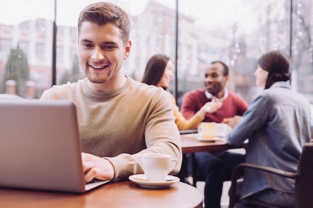 Atractivo hombre agradable lindo escribiendo en la computadora portátil mientras disfruta de un café y emerge en el mundo virtual