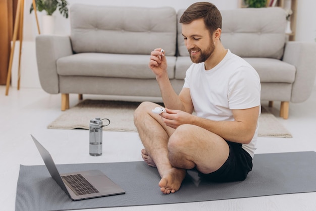 Atractivo hombre barbudo que sostiene auriculares inalámbricos cerca de la computadora portátil y comienza el ejercicio matutino o el yoga