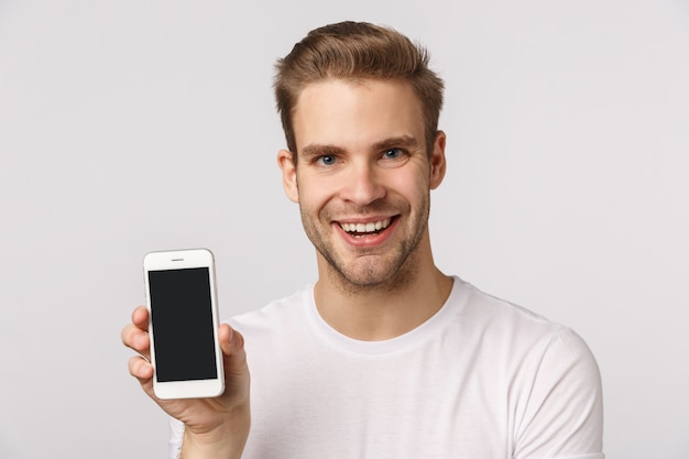 Atractivo hombre barbudo rubio en camiseta blanca mostrando smartphone