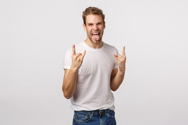 Atractivo hombre barbudo rubio en camiseta blanca que muestra signo de heavy metal