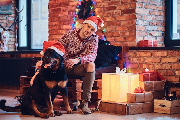 Atractivo hombre hipster barbudo con su perro Rottweiler en una habitación con decoración navideña.