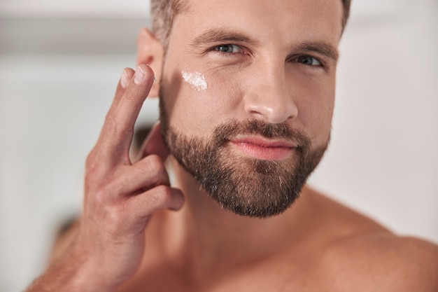 Atractivo hombre maduro feliz con barba elegante aplica crema sobre la piel debajo de los ojos en primer plano de baño ligero