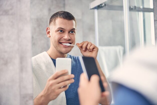 Atractivo hombre morena limpiando los dientes del espejo mientras sostiene el móvil en la mano