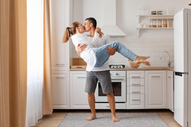 Atractivo hombre y mujer de familia divirtiéndose en la cocina celebrando la reubicación del tipo sosteniendo a su novia o esposa y besándose expresando sentimientos románticos