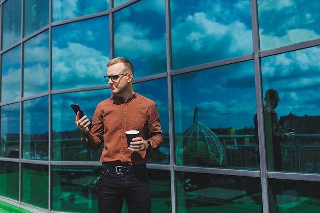 Atractivo hombre de negocios de moda con una elegante camisa y pantalones hablando por teléfono cerca del edificio de oficinas y bebiendo café oficina moderna de vidrio