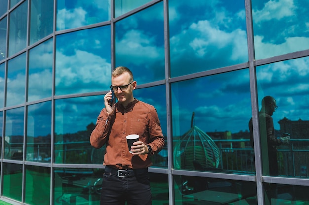 Atractivo hombre de negocios de moda con una elegante camisa y pantalones hablando por teléfono cerca del edificio de oficinas y bebiendo café oficina moderna de vidrio