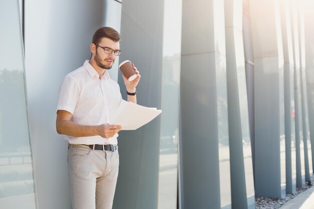 Atractivo hombre de negocios o abogado con anteojos está revisando documentos de contrato mientras está parado cerca de un edificio de oficinas moderno, copiando espacio. Concepto de negocio y éxito