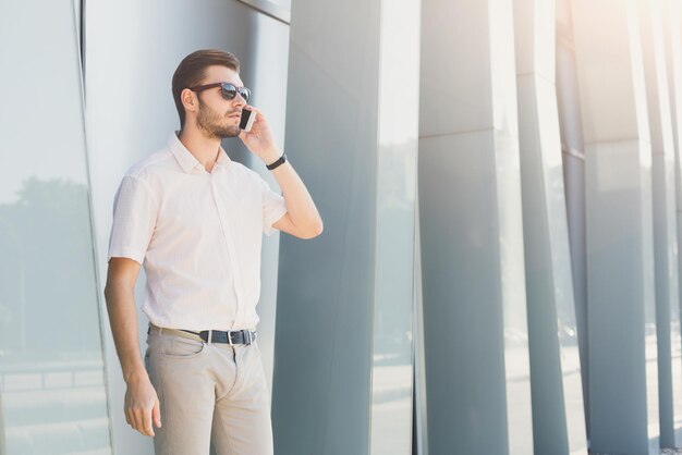 Atractivo hombre de negocios o abogado con gafas de sol está hablando por teléfono celular y mirando hacia otro lado cerca de un edificio de oficinas, copiando espacio. Concepto de negocio, tecnología y comunicación.