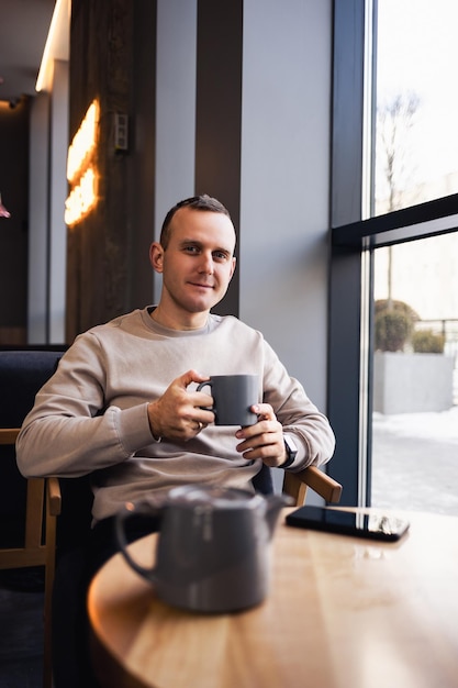 Un atractivo hombre solitario se sienta en una mesa de café, bebe té caliente fragante. Un tipo sonriente con ropa informal se sienta en una cafetería y bebe café.