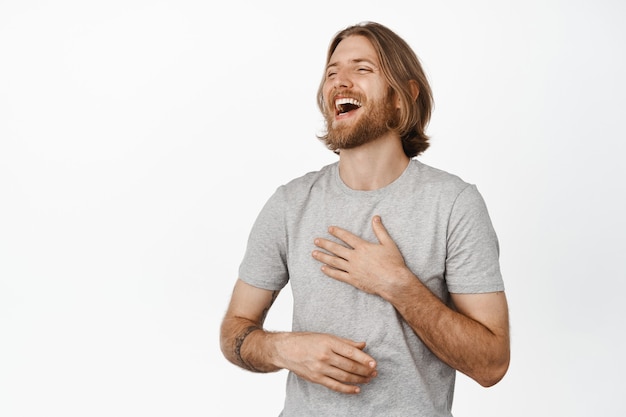 Atractivo hombre sueco con elegante cabello y barba se ríe de la divertida historia de un amigo con las manos cruzadas y los ojos cerrados.