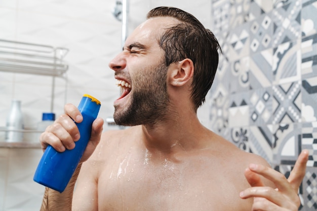 Atractivo joven alegre cantando mientras se lava en la ducha, sosteniendo la botella de champú