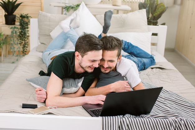 Atractivo joven apoyado en su novio en la cama, mirando un portátil. Pareja gay acostado en la cama y usando la computadora portátil en casa