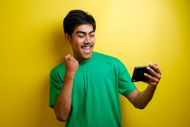 Atractivo joven asiático tiene buenas noticias en su teléfono, sonriendo feliz riendo expresión ganadora contra el fondo amarillo