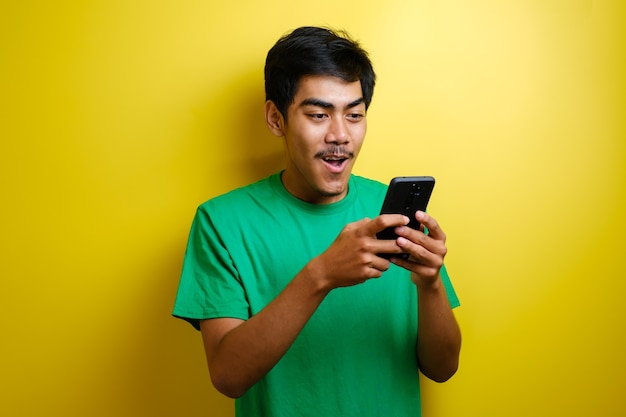Atractivo joven asiático tiene buenas noticias en su teléfono, sonriendo feliz riendo expresión ganadora contra el fondo amarillo