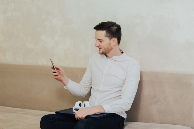 Atractivo joven barbudo con camisa blanca sentado en un sofá en la sala de estar, mediante teléfono móvil.