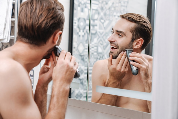 Atractivo joven sin camisa afeitado sonriente mientras está de pie en el espejo del baño, con maquinilla de afeitar eléctrica