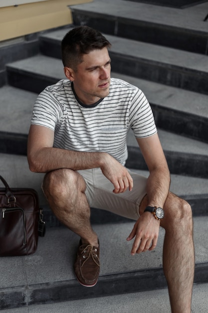 Atractivo joven con una elegante camiseta a rayas en elegantes pantalones cortos en zapatos de gamuza con una bolsa de cuero se sienta en una piedra vintage