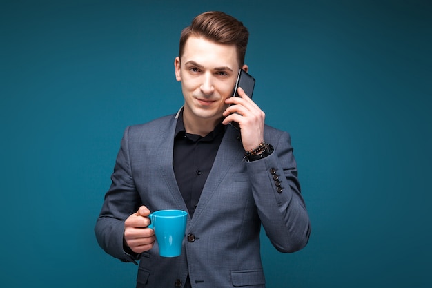 Atractivo joven empresario en chaqueta gris y camisa negra sostenga la taza azul y hable por teléfono