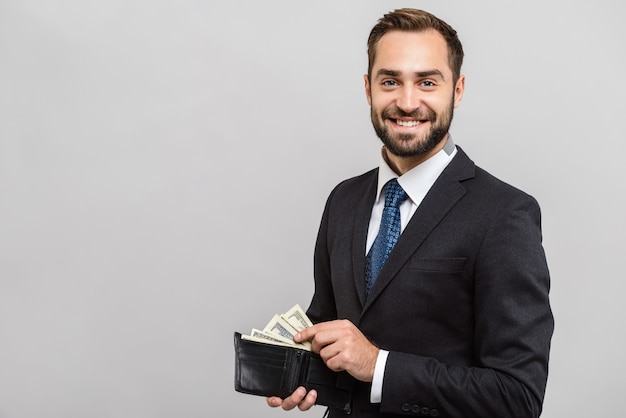 Atractivo joven empresario feliz vistiendo traje que se encuentran aisladas sobre pared gris, mostrando la billetera llena de billetes de dinero