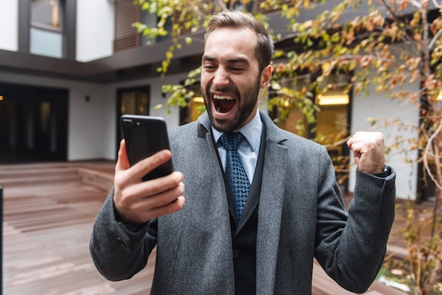 Atractivo joven empresario vistiendo traje caminando al aire libre, mediante teléfono móvil, celebrando