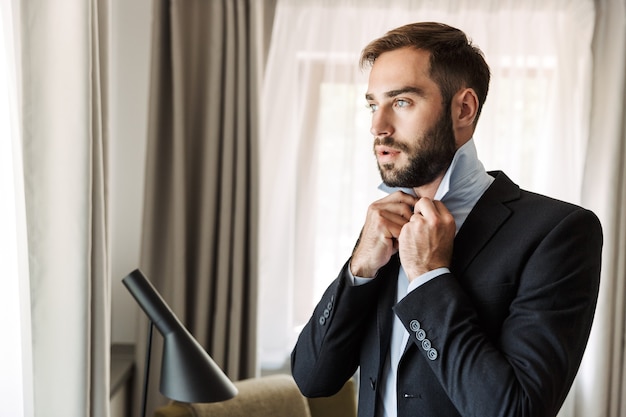 Atractivo joven empresario vistiendo traje de pie en la habitación del hotel, vistiéndose