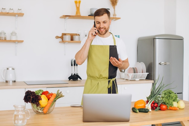 Atractivo joven está cocinando en la cocina con una computadora portátil en la mesa hablando en un teléfono inteligente