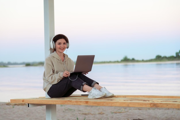 Atractivo joven freelancer trabajando en la playa con auriculares y laptop sonriendo