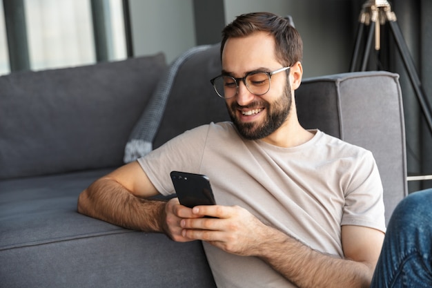 Atractivo joven inteligente sentado en el piso de la sala de estar, mediante teléfono móvil