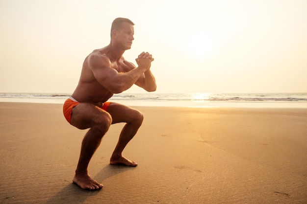 Atractivo joven modelo masculino musculoso haciendo el ejercicio de estiramiento calentarse en la playa summersexy atleta abs y cuerpo perfecto en el paraíso tropical puesta de sol en la costa