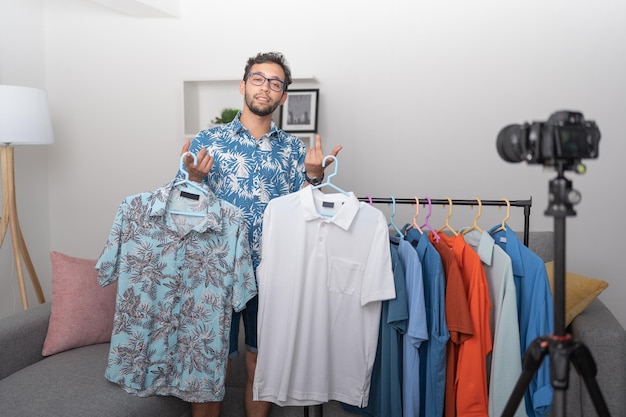 Atractivo joven mostrando los nuevos modelos y colores de camisetas que tiene mientras graba un video blog