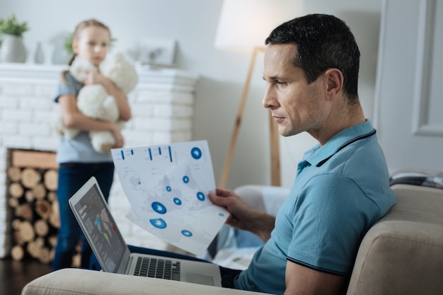 Foto atractivo joven de pelo oscuro serio sosteniendo papeles y mirando la computadora portátil mientras su hija de pie con su juguete y mirándolo