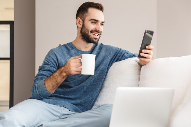 Atractivo joven relajándose en un sofá en casa, mediante teléfono móvil, tomando café