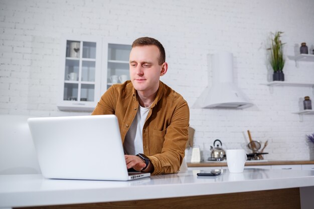 Un atractivo joven en ropa casual sentado en la cocina usando una computadora portátil. Trabaje desde casa, flujo de trabajo remoto.