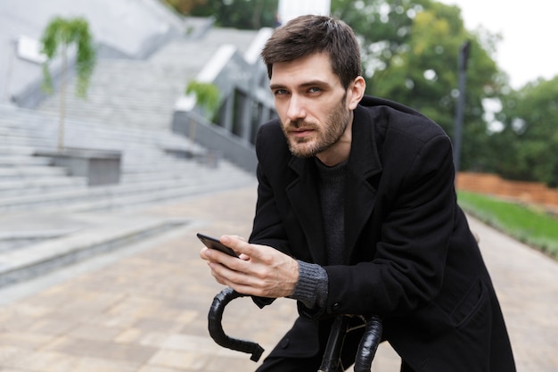 Atractivo joven vestido con abrigo apoyado en una bicicleta en una calle, teléfono móvil usnig