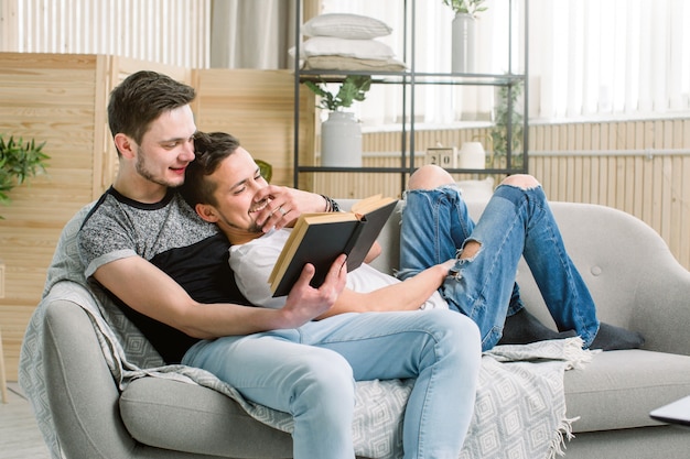 Atractivo libro de lectura de pareja gay en el sofá en casa en la sala de estar