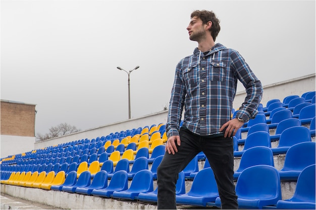 Atractivo modelo deportivo joven en camisa azul sentado en los asientos del estadio azul después del entrenamiento mirando al campo